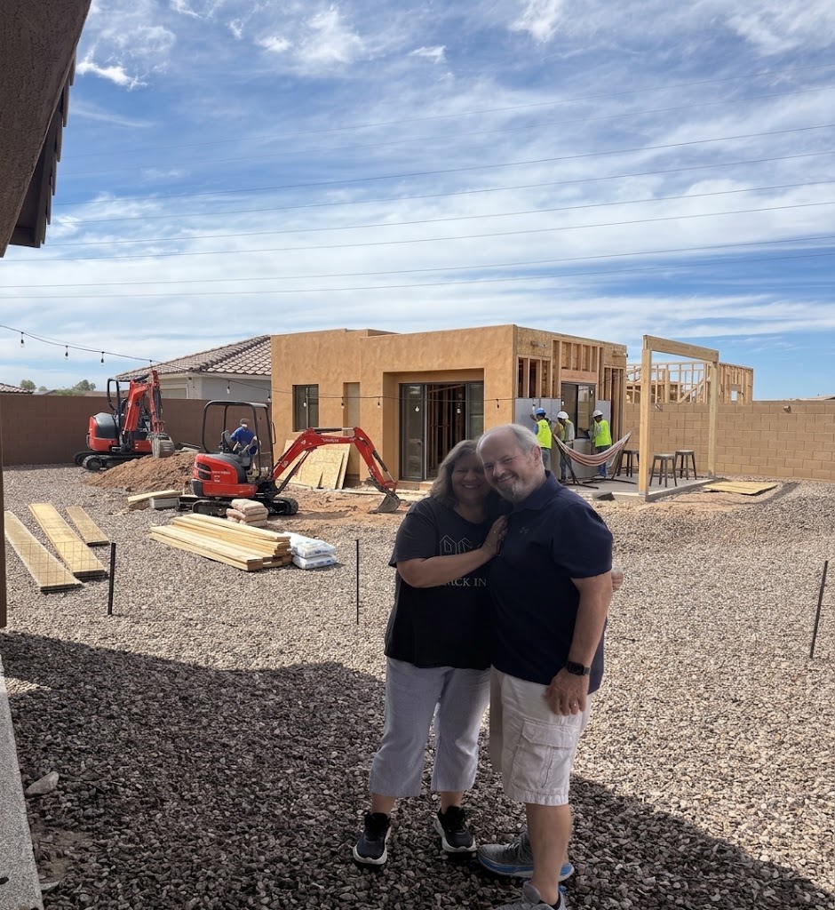 A couple standing together in front of a casita under construction, with framing and a construction crew in the background