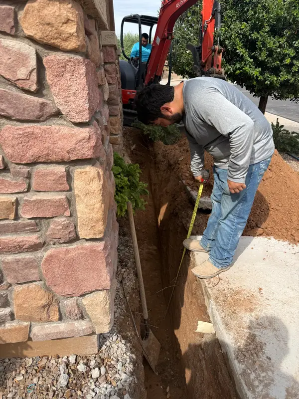 Maricopa Casitas crew member measuring the depth of a utility trench beside a stone wall, with a Kubota mini excavator behind him